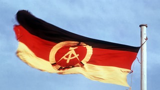 Die Flagge der Deutschen Demokratischen Republik: schwarz-rot-gold mit Hammer und Zirkel.