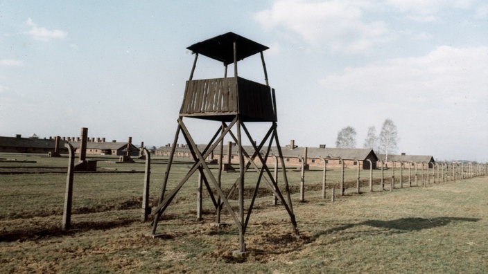 Ein hölzerner Wachturm vor einem Stacheldrahtzaun. Im Hintergrund stehen mehrere flache lange Ziegelbauten.