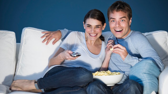 Ein Pärchen sitzt auf einem Sofa, isst Popcorn und schaut fern.