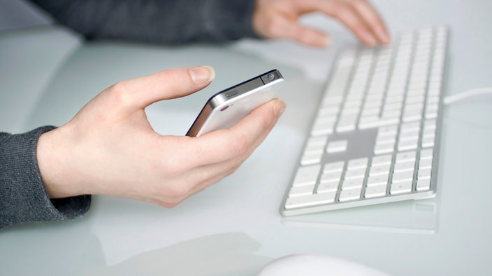 Eine Person sitzt am Computer und hält gleichzeitig das Handy in der Hand.