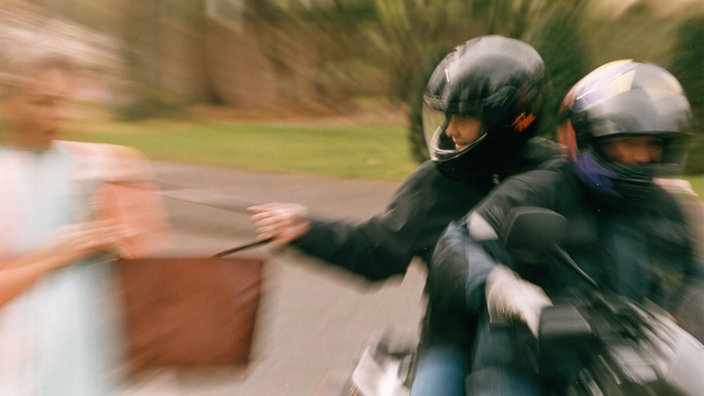 Zwei Männer auf einem Roller klauen eine Tasche einer Frau.