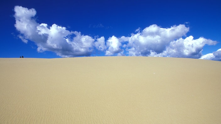 Eine große Dünenlandschaft im Vordergrund, weit hinten zwei winzige Köpfe.