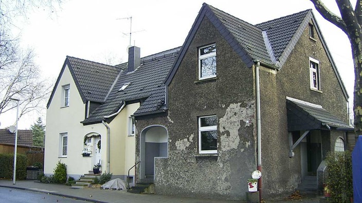 Eineinhalbgeschossiges Doppelhaus mit zwei Eingängen in der Mitte. Die linke Seite des Hauses ist renoviert. Die Fassade ist frisch gestrichen. An der Tür hängt ein Blumenkranz. Die rechte Seite ist nicht renoviert. Der dunkle Putz blättert ab.