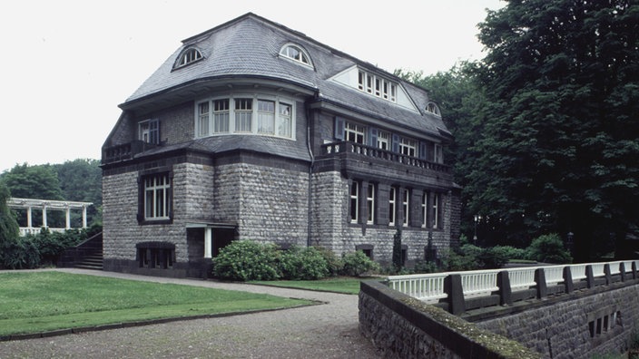 Eine schiefergedeckte Villa aus grauen Bruchsteinen in einer parkähnlichen Landschaft.