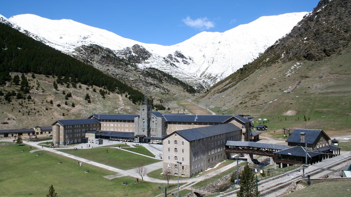 Blick auf ein großes, kastenförmiges Gebäude mit zwei Seitenflügeln, das von schneebedeckten Bergen umgeben ist.