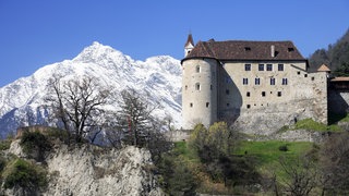 Im Hintergrund ein schneebedeckter Berg, im Vordergrund eine mittelalterliche Burg auf einem Felssporn.