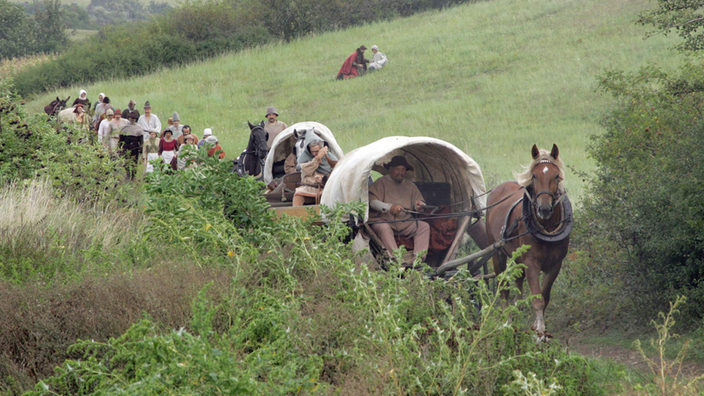 Zwei Planwagen und mehrere Menschen zu Fuß in einer grünen Landschaft.