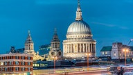 Stadtpanorama in schönem Abendlicht. Vorne sieht man den Fluss Themse, dahinter eine Brücke, hinter ihr den großen Kuppelbau der Kirche St. Paul's Cathedral, andere größere Häuser sowie zwei weitere Kirchtürme