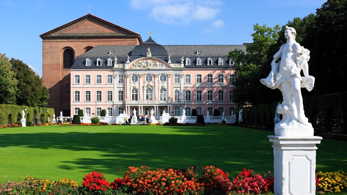 Ein Blick bei herrlichem Sommerwetter vom Schlosspark auf die prachtvolle Rokoko-Fassade des kurfürstlichen Palais. Links angrenzend die Basilika aus der Römerzeit. Im Vordergrund eine Statue und Blumenbeet.