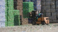 Kombrimierte Berge aus Plastikmüll, davor fährt ein Staplerfahrer.