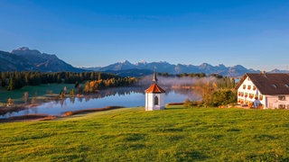 Blick auf ein Bauernhaus und eine Kapelle, die an einem See im Allgäu liegen. Im Hintergrund die Silhouette vom Hochgebirge.