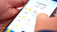 Zwei Hände halten ein Smartphone, auf dem eine Wetter-App geöffnet ist.